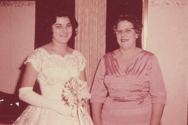 Two women, one in a white gown holding flowers, the other in a pink dress smile and look at the camera.