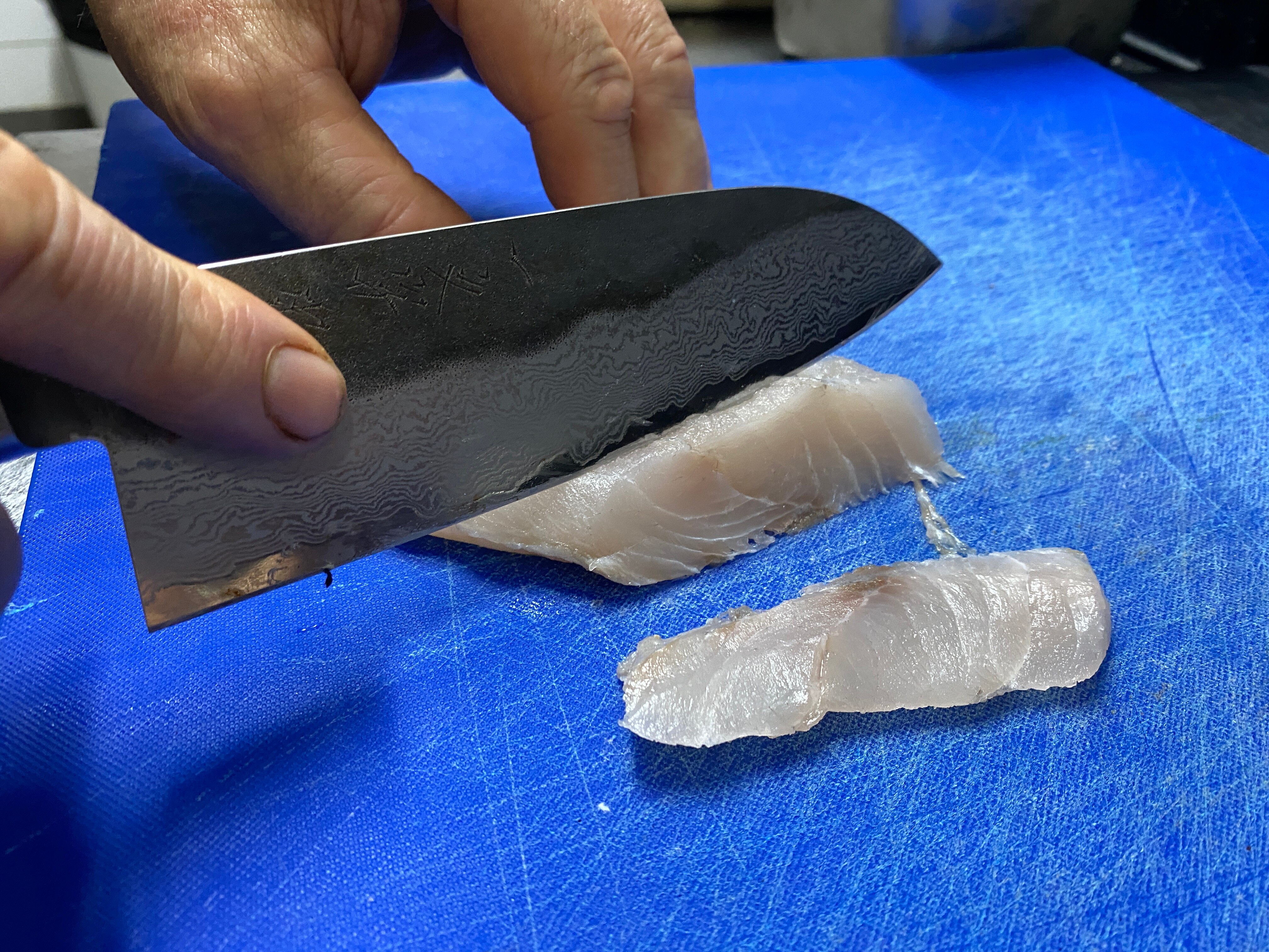 A close up of a knife cutting through a barramundi peice on a blue chopping board.