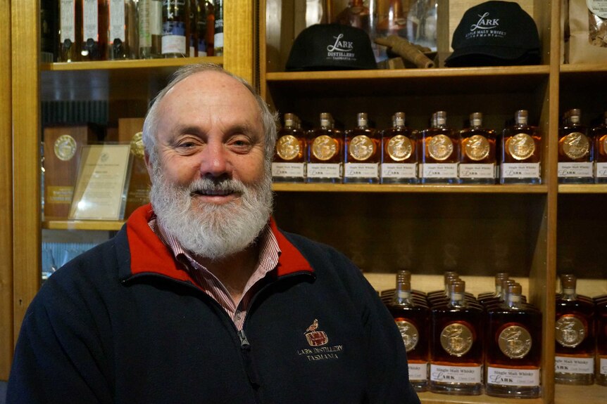 Smiling older man, grey beard, moustache, balding, wears red-collar jumper, shirt, stands in front of whisky bottles.