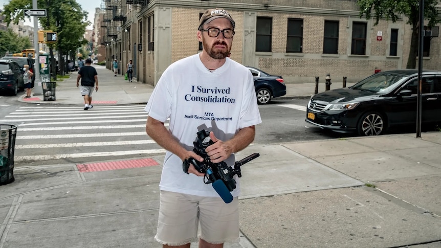 A man on NYC streets hokding a camera, in a white t-shirt that reads "I survived consolidation. MTA Audit Services Department".
