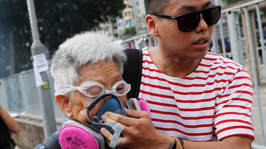A young man holds a breathing mask to an elderly's woman's face as they move away from tear gas.