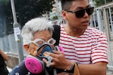 A young man holds a breathing mask to an elderly's woman's face as they move away from tear gas.