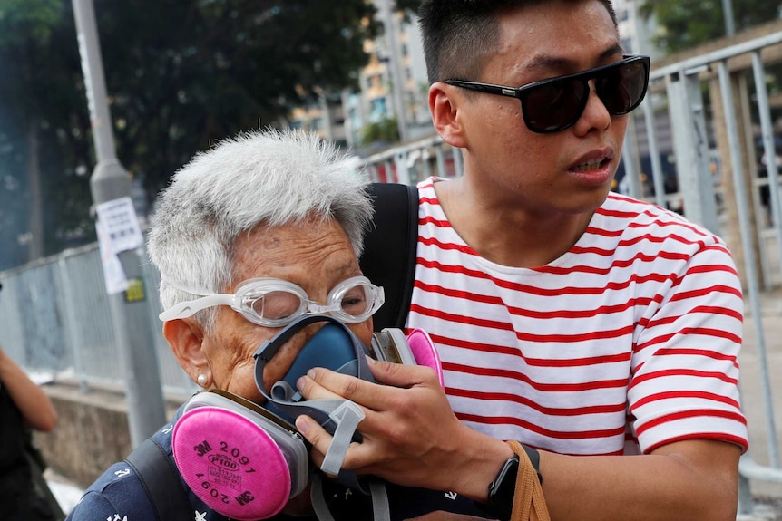 A young man holds a breathing mask to an elderly's woman's face as they move away from tear gas.