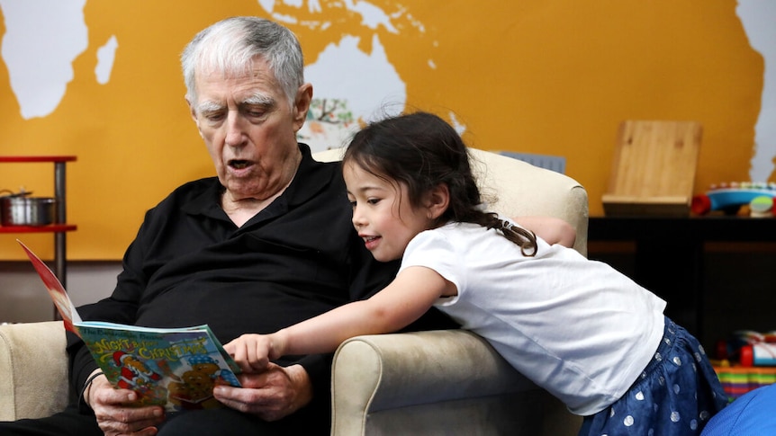 An older man reading a book next to a dark-haired four year old girl, pointing at the book.