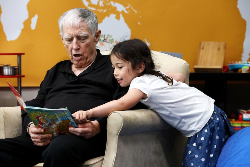 An older man reading a book next to a dark-haired four year old girl, pointing at the book.