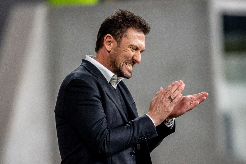 Tony Popovic claps with his eyes closed and mouth clenched wearing a black suit jacket