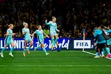 A women's soccer team wearing light blue uniforms runs to the sideline celebrating