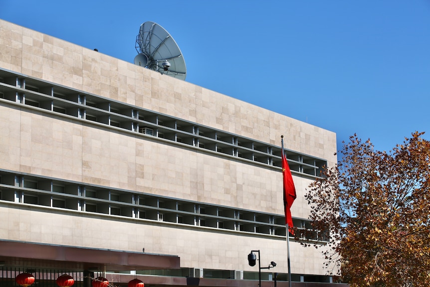 A three storey building behind a fence. On top sits a satellite, in front is a Chinese flag.