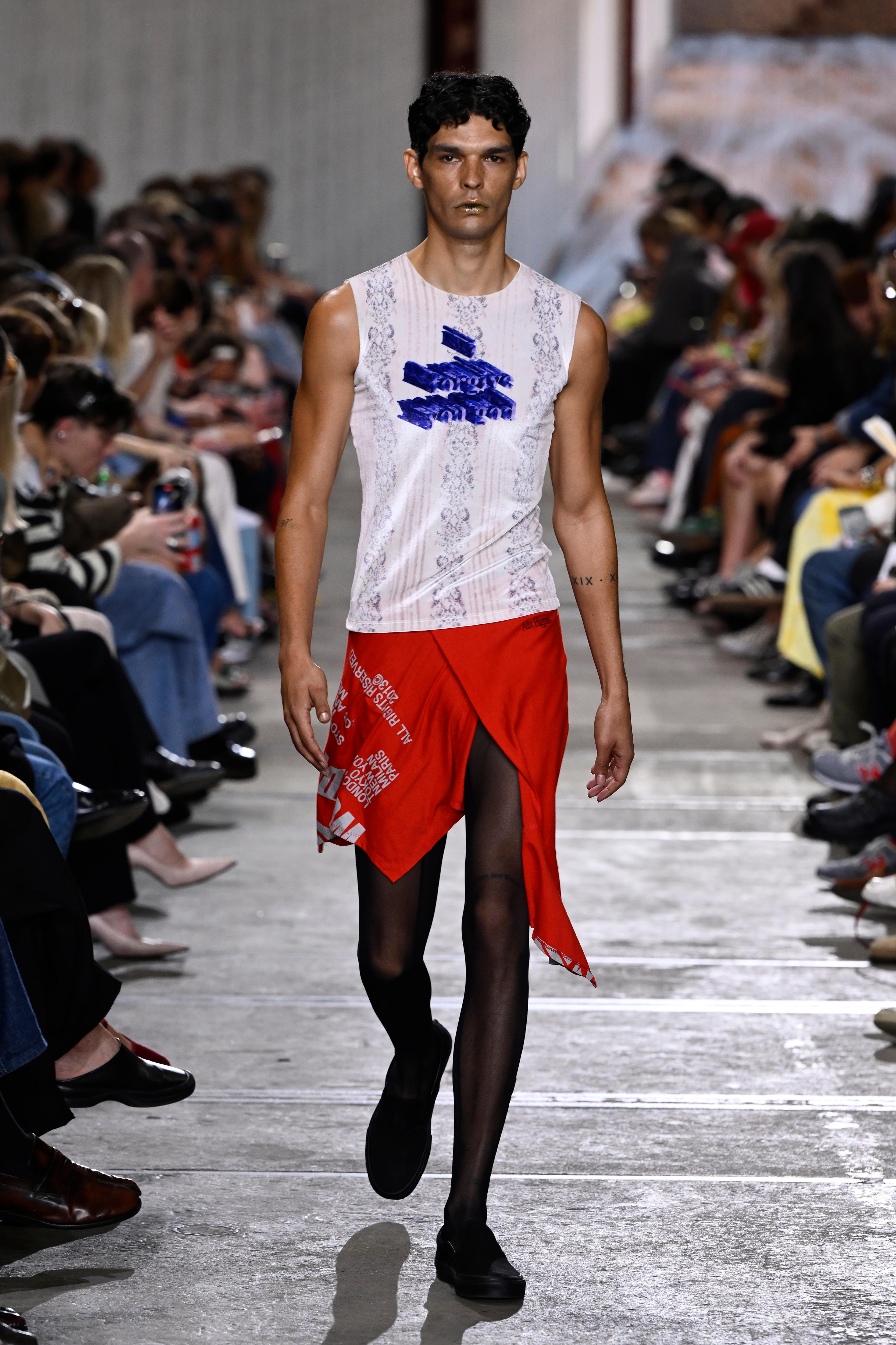 A man walks Higgins 2025 resort runway in a short red skirt and a tight singlet, the show's title printed over a floral print..