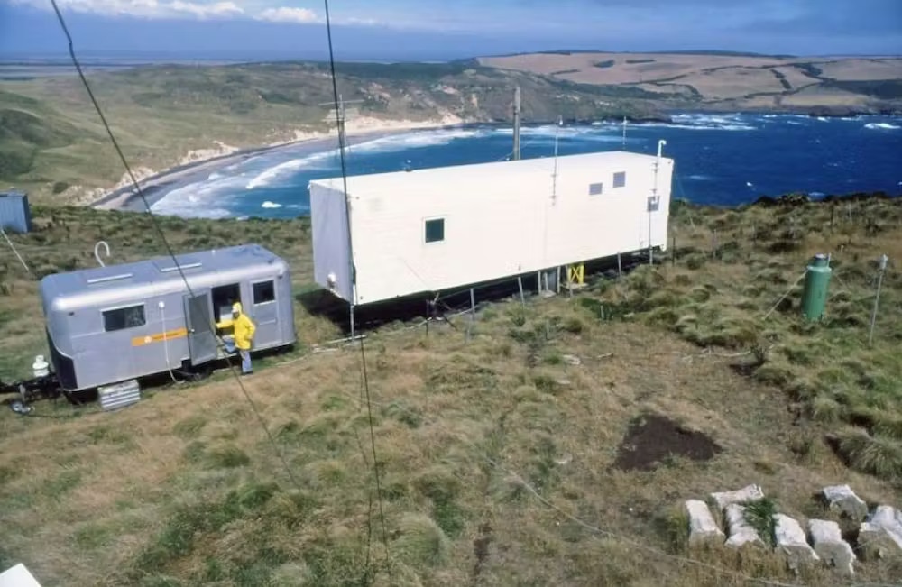 A silver caravan sits next to a white rectangular structure on a cliff overlooking blue water.