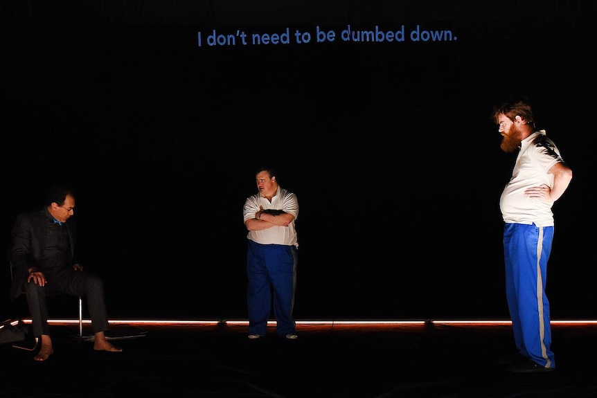 Black-backed theatre stage with three figures, and text across back wall saying "I don't need to be dumbed down".