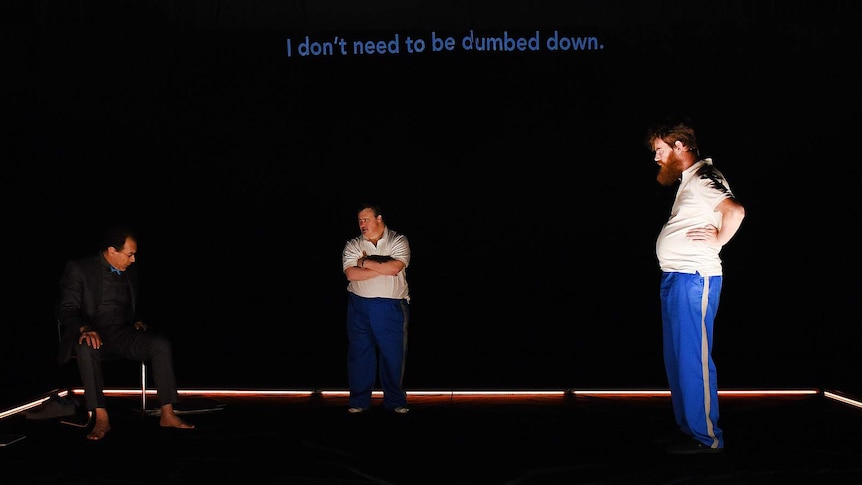 Black-backed theatre stage with three figures, and text across back wall saying "I don't need to be dumbed down".