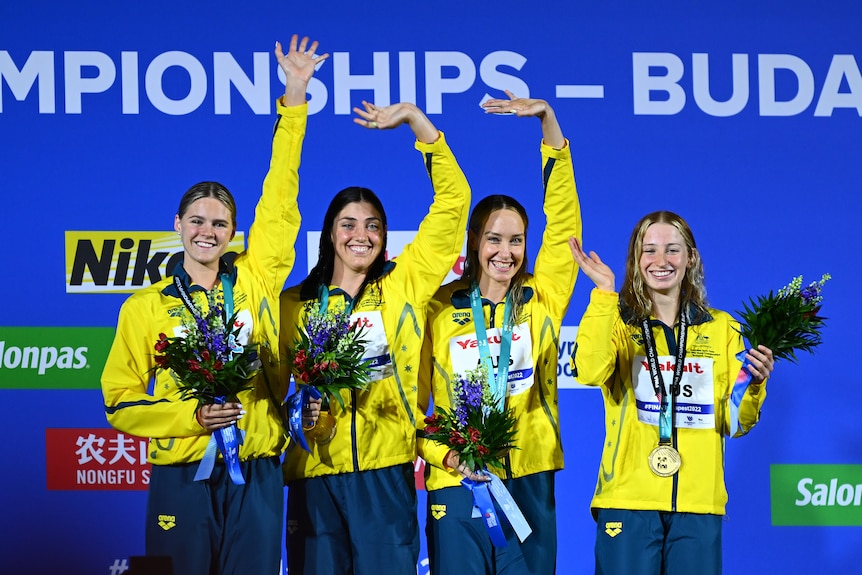 Four Australian female swimmers smile and wave from the podium with gold medals around their necks.