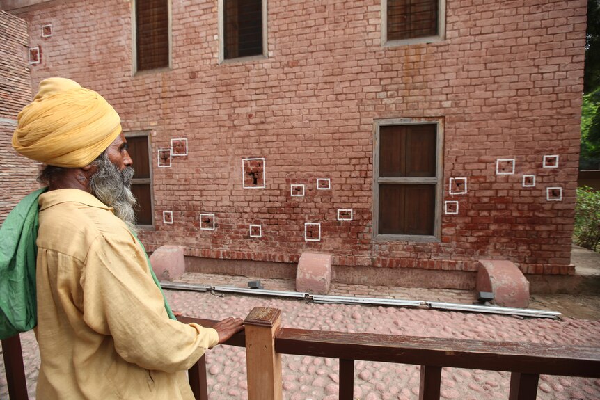 A man dressed in a yellow turban and outfit looks at a wall sprinkled with bullet holes.