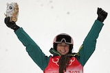 A smiling woman in green ski gear holds a gold statue above her head