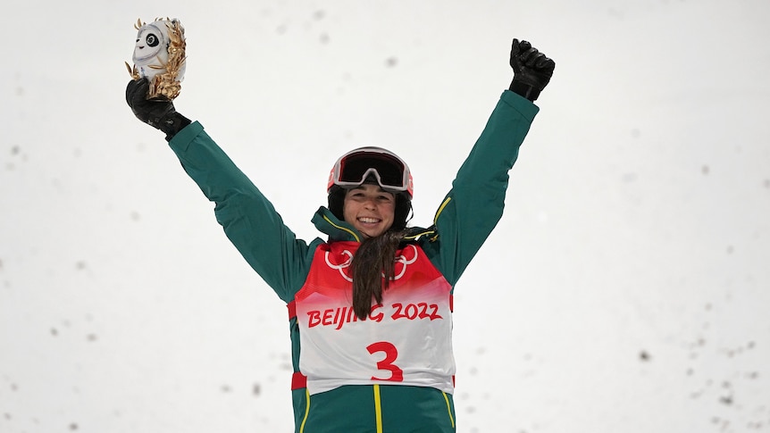 A smiling woman in green ski gear holds a gold statue above her head