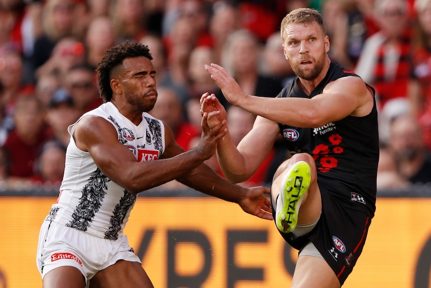 An Essendon AFL player kicks the ball while under defensive pressure from a Collingwood opponent.