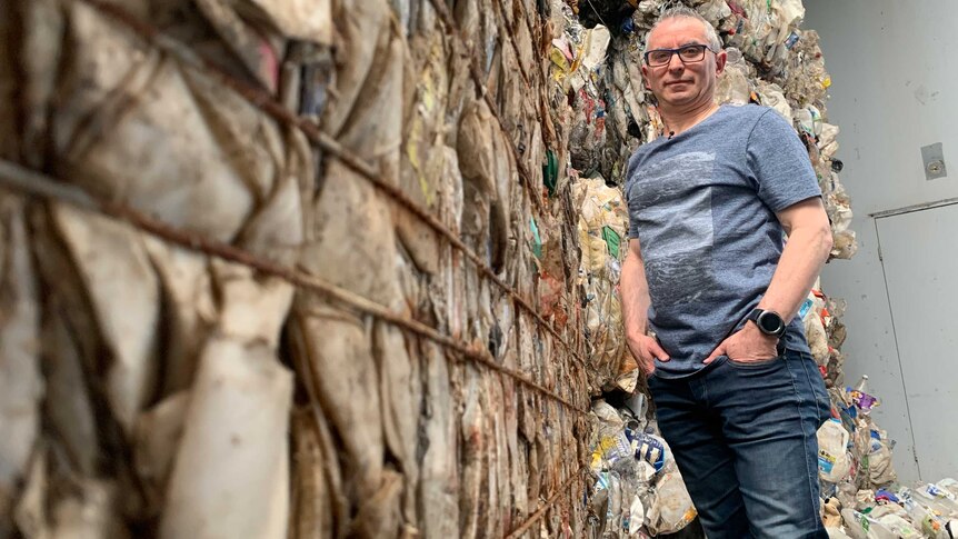 A picture of a man with grey hair and glasses, standing in jeans and a t-shirt and surrounded by piles of rubbish.