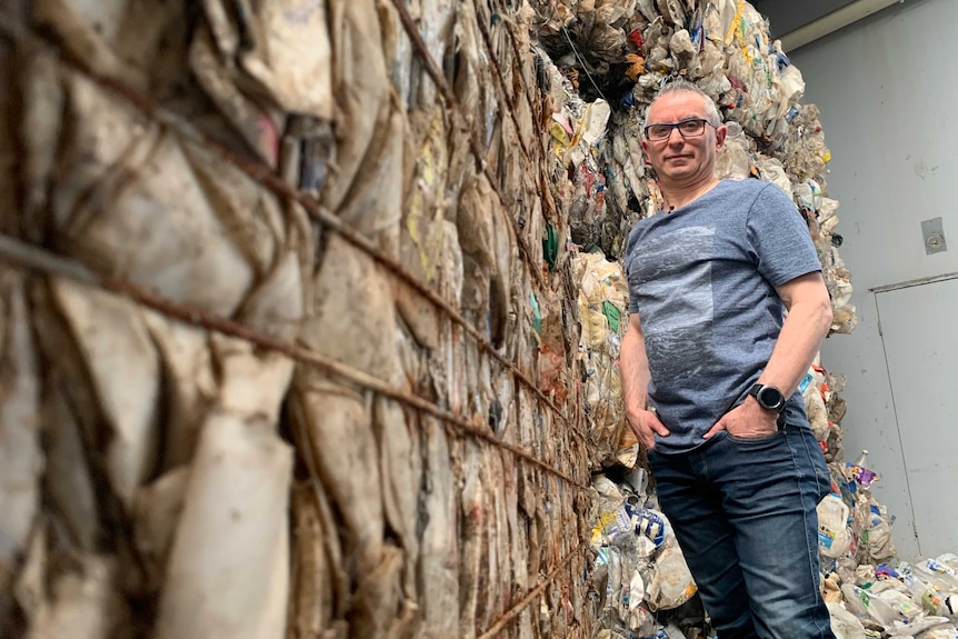 A picture of a man with grey hair and glasses, standing in jeans and a t-shirt and surrounded by piles of rubbish.