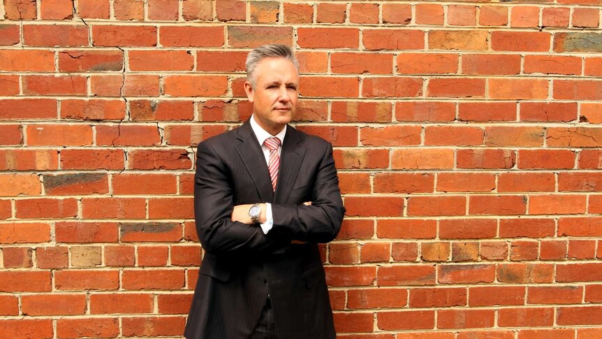 A man in a suit stands against a brick wall.