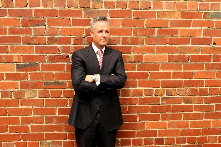 A man in a suit stands against a brick wall.