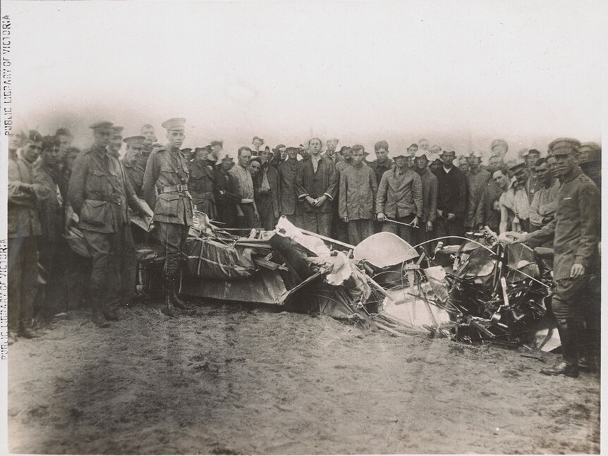 The crumpled remains of a plane's fuselage on sand, surrounded by men in uniform.