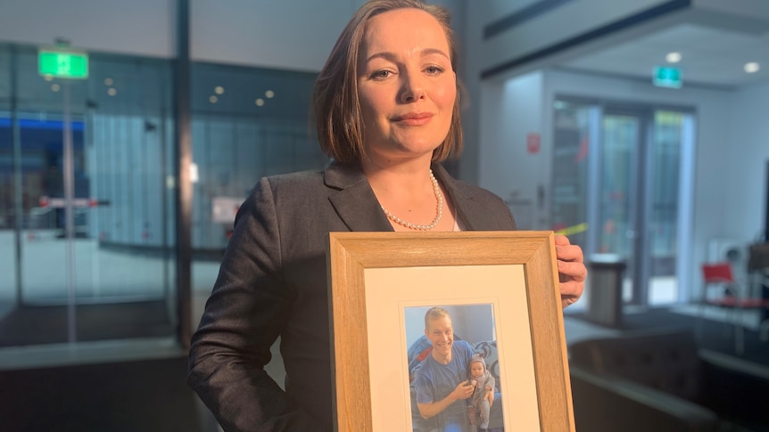 A woman holds a wooden frame, containing a photo of a smiling father holding a baby girl.