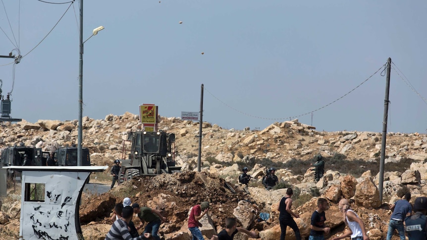 Palestinian youths throw stones at armed  Israeli soldiers across  a mound of rocks being bulldozed across a road.
