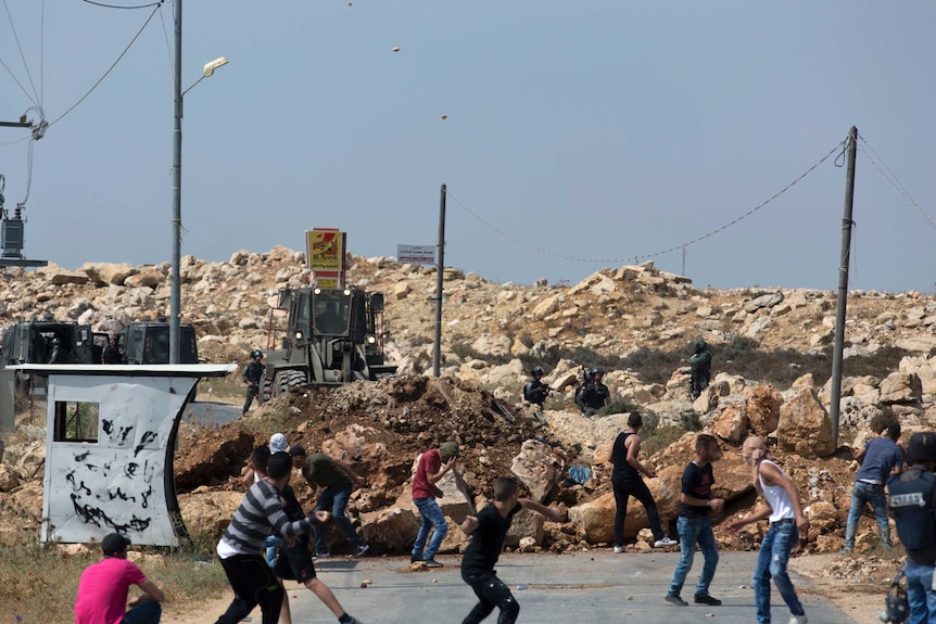 Palestinian youths throw stones at armed  Israeli soldiers across  a mound of rocks being bulldozed across a road.