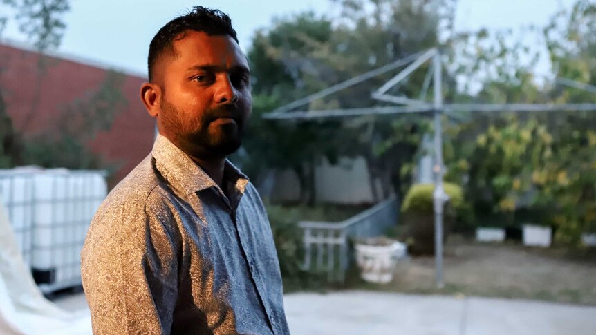 Man wearing collared shirt with beard standing outside in backyard with Hills Hoist and brick wall behind him.