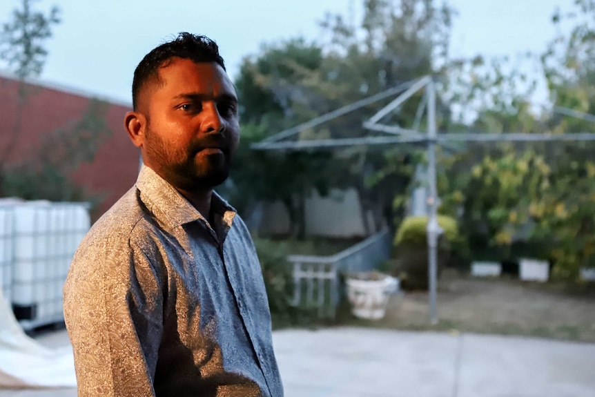 Man wearing collared shirt with beard standing outside in backyard with Hills Hoist and brick wall behind him.