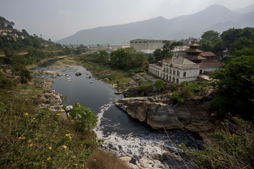 The polluted Bagmati River flows  with greenery, houses and mountains surrounding it on a grey-sky day 