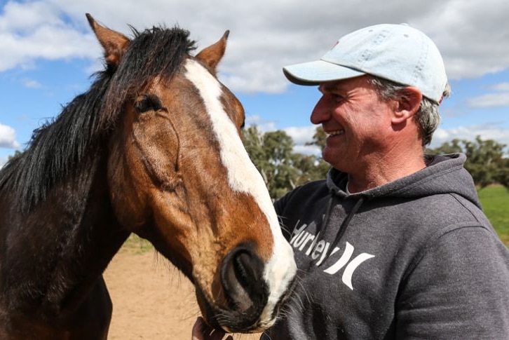 A smiling man in a cap and hoodie standing with a racehorse.