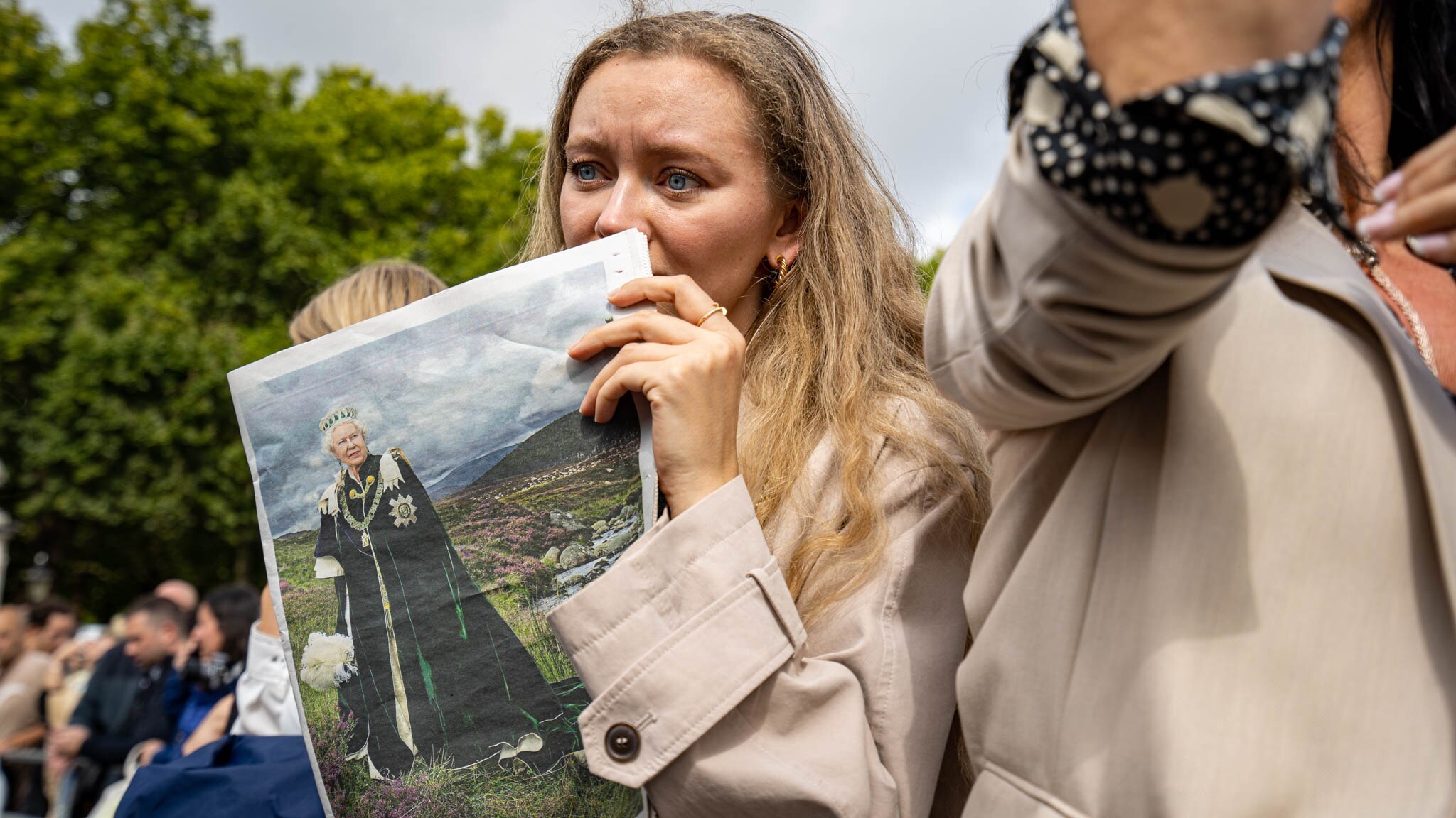 A woman with long hair wearing a trench coat holds a newspaper to her face, looking emotional 