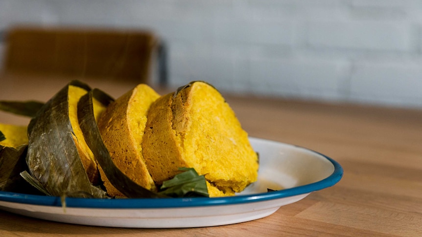 Four slices of yellow bread unwrapped from banana leaf on a blue-rimmed white plate set on a wooden cafe table.