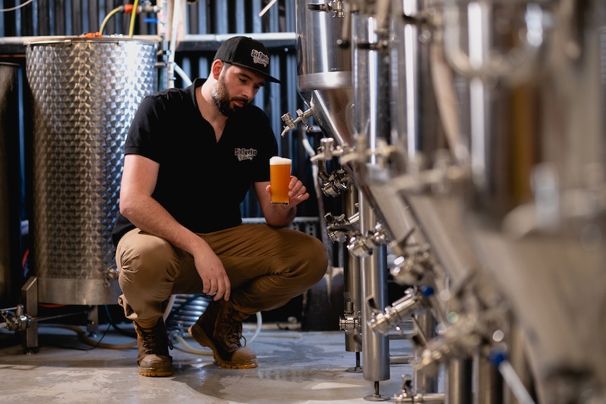 A man studies a beer that is tapped from a tap next to stainless steel tanks