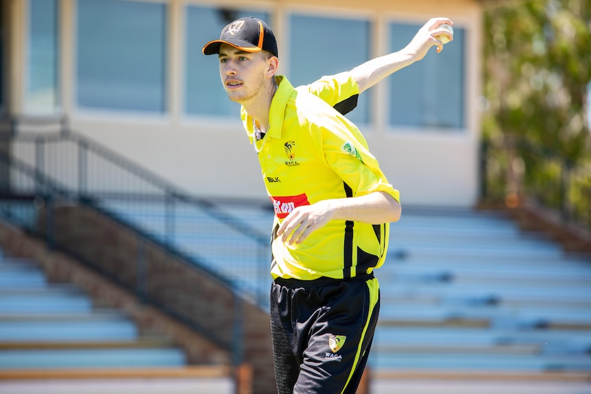 A man in a yellow shirt bowls a cricket ball underarm.