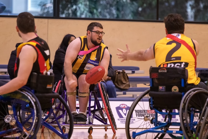 A Wheelchair AFL match, in focus is a man in yellow and brown footy jersey with a ball he's about to pass