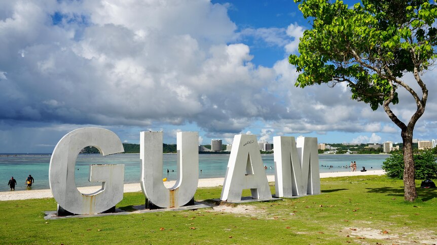 Large metal letters spelling GUAM sit on grass next to a tree by the beachfront