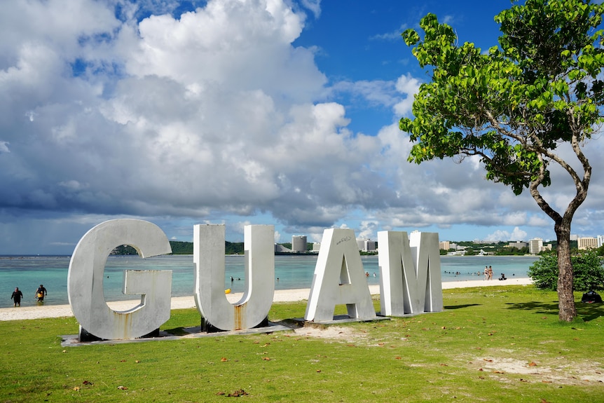 Large metal letters spelling GUAM sit on grass next to a tree by the beachfront