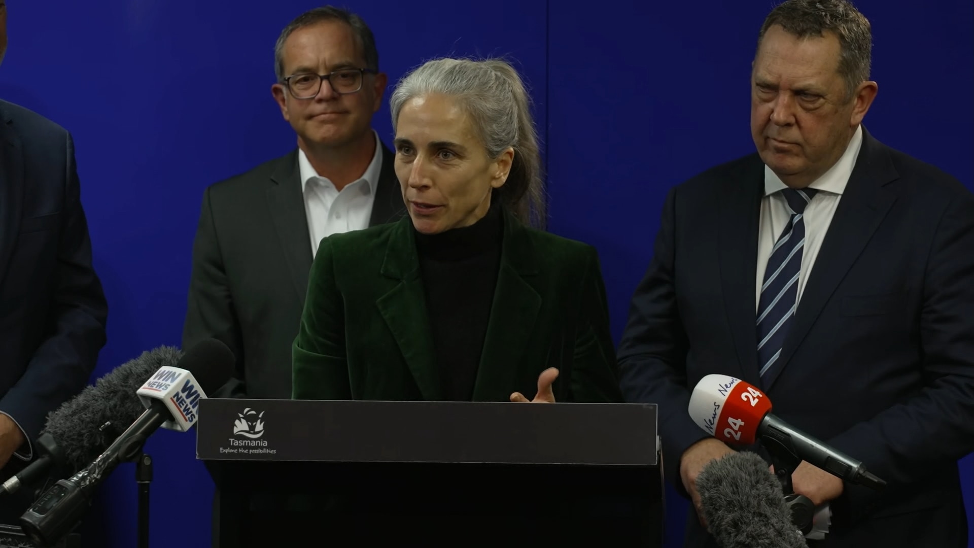 a woman with a grey ponytail speaks at a lecturn, flanked by two men.