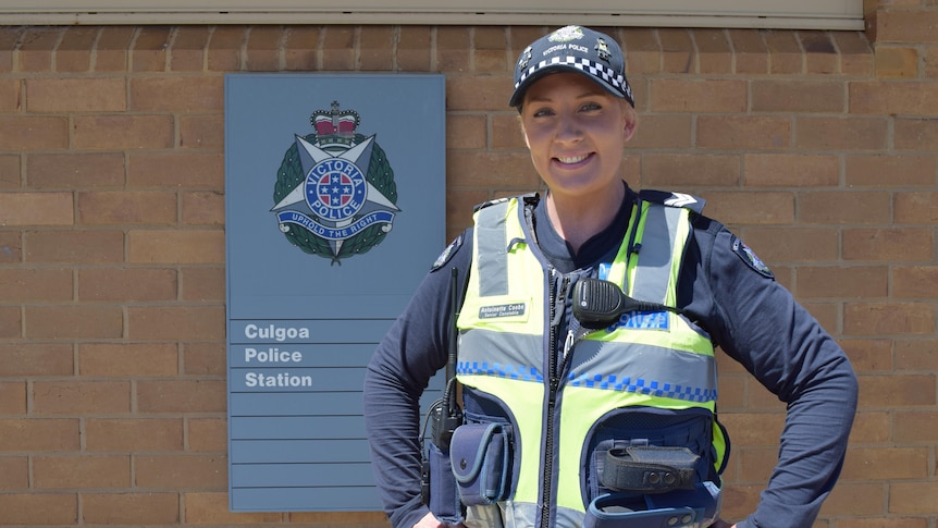A policewoman smiles in front of a blue sign on a brick wall reading Culgoa police station.