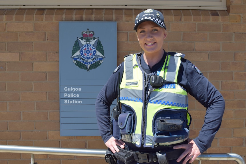 A policewoman smiles in front of a blue sign on a brick wall reading Culgoa police station