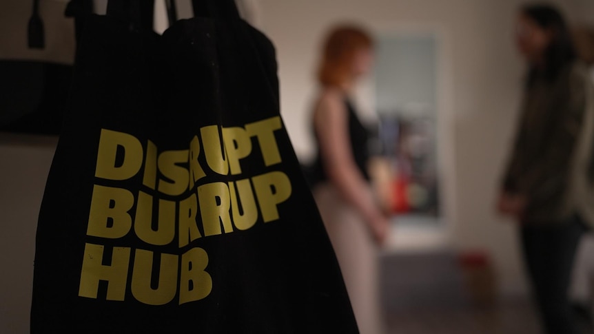 A bag saying 'DISRUPT BURRUP HUB' hangs as two people talk in the background.