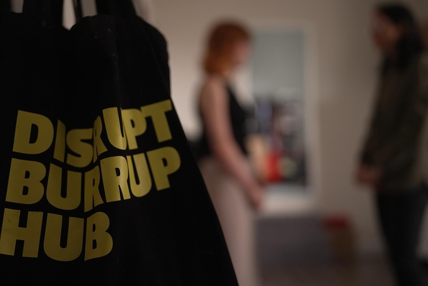 A bag saying 'DISRUPT BURRUP HUB' hangs as two people talk in the background.