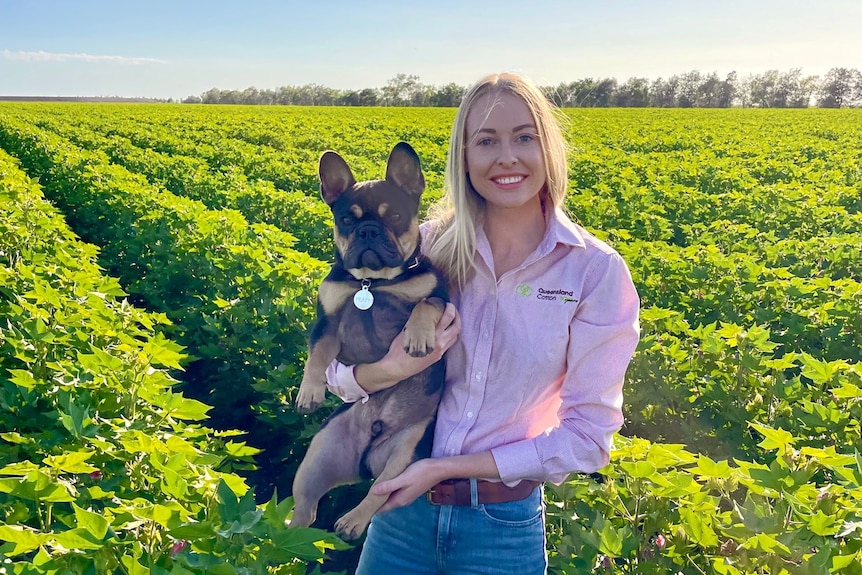 Jess Strauch stands in a field holding a dog, trees are visible in the background.