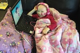 Charlotte Jones wearing headphones and laying down under blanket watching cartoon on laptop.