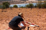 A female scientists wearing a brown hat crouches on dirt ground at a dig site near the River Murray.