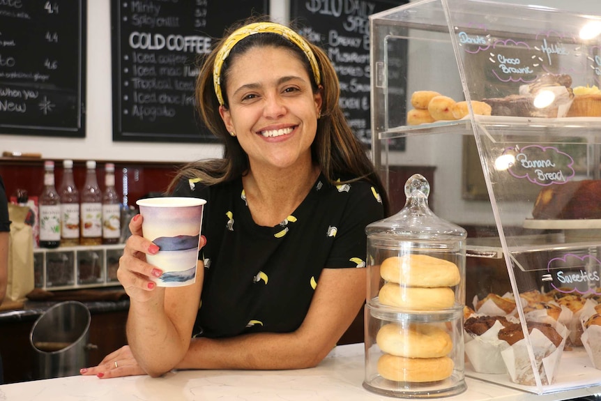 A woman with a black top and yellow hair band on stands behind a counter in a coffee shop holding a cup of coffee and smiling.