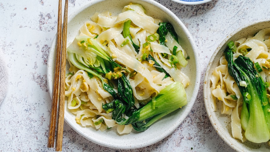 A pair of chopsticks rests on the edge of a bowl filled with noodles and bok choy.
